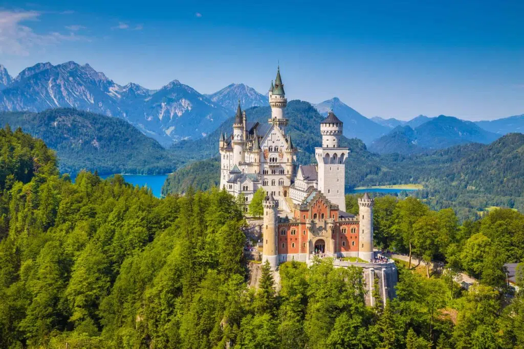 Vista aérea do Castelo de Neuschwanstein, com árvores em volta e um belo lago azul atrás.