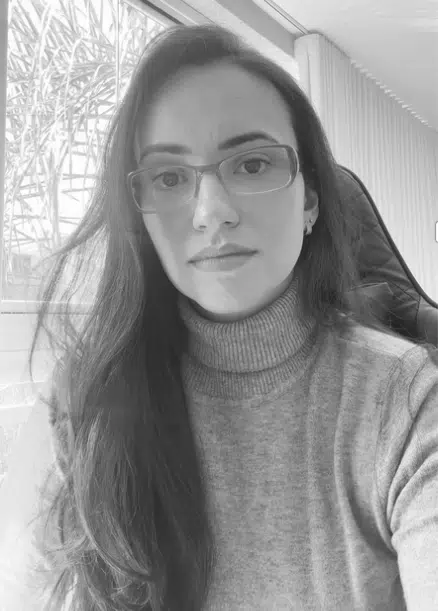 Gabriella-Vilafranca Mulher de óculos usando um suéter de gola alta, com um olhar pensativo e elegante.