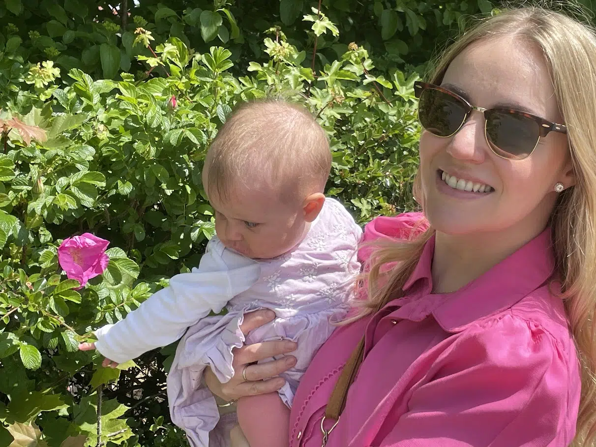 Mulher usando óculos escuros, segurando um bebê com uma camiseta rosa.