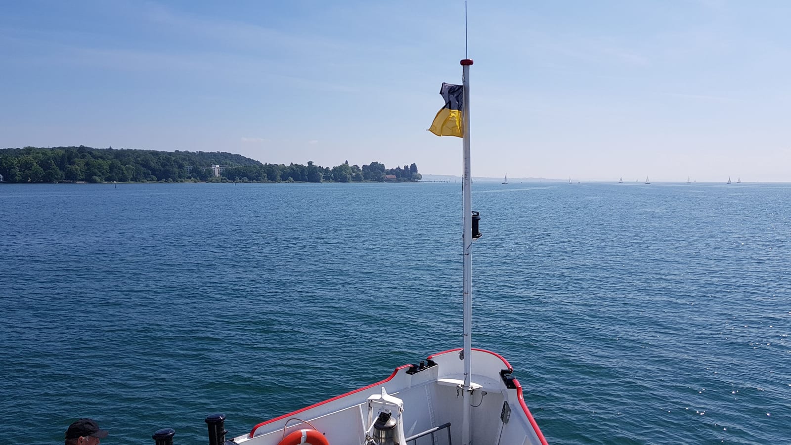 Proa de uma balsa no Lago de Constança, com a bandeira no mastro e a margem ao fundo