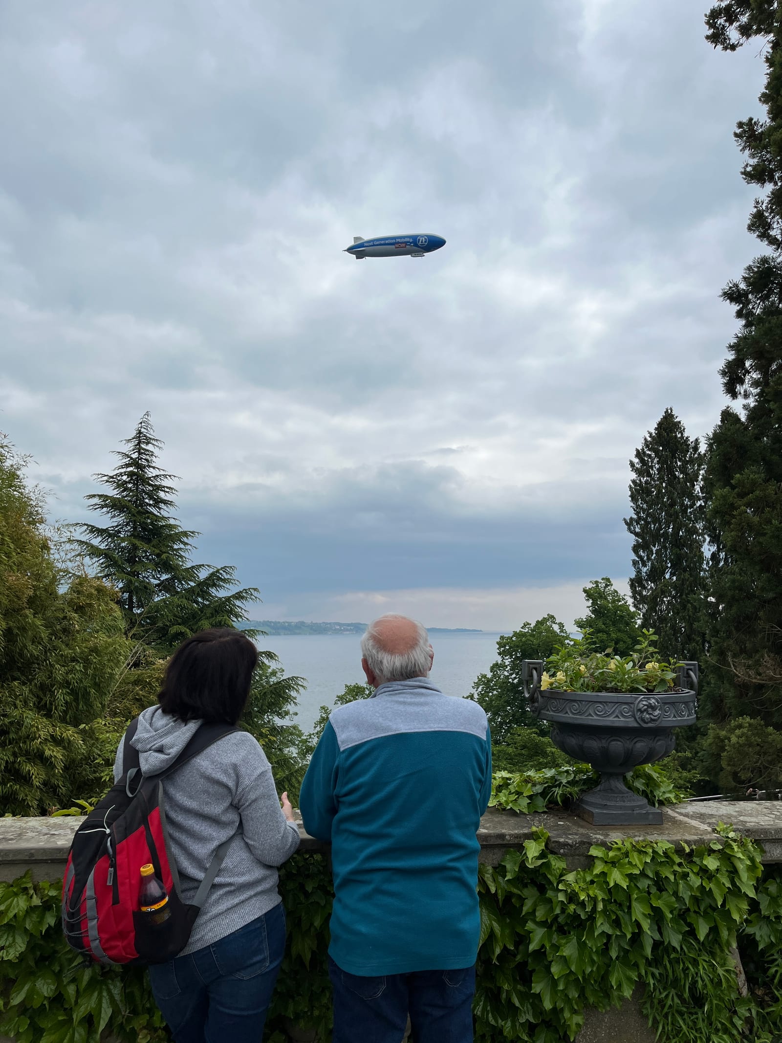 Visitantes contemplam o Lago de Constança a partir de um mirante, com um Zeppelin ao longe