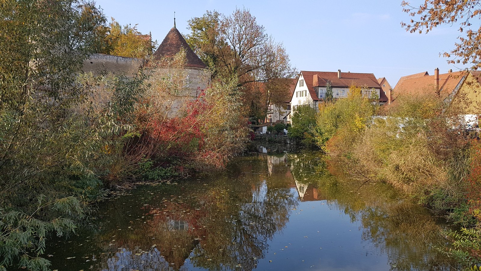 Muralha e torre de Dinkelsbühl refletidas na água, com árvores coloridas de outono