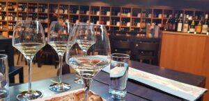 Três taças de vinho branco alemão numa mesa de degustação, com um mapa da região e prateleiras de garrafas ao fundo