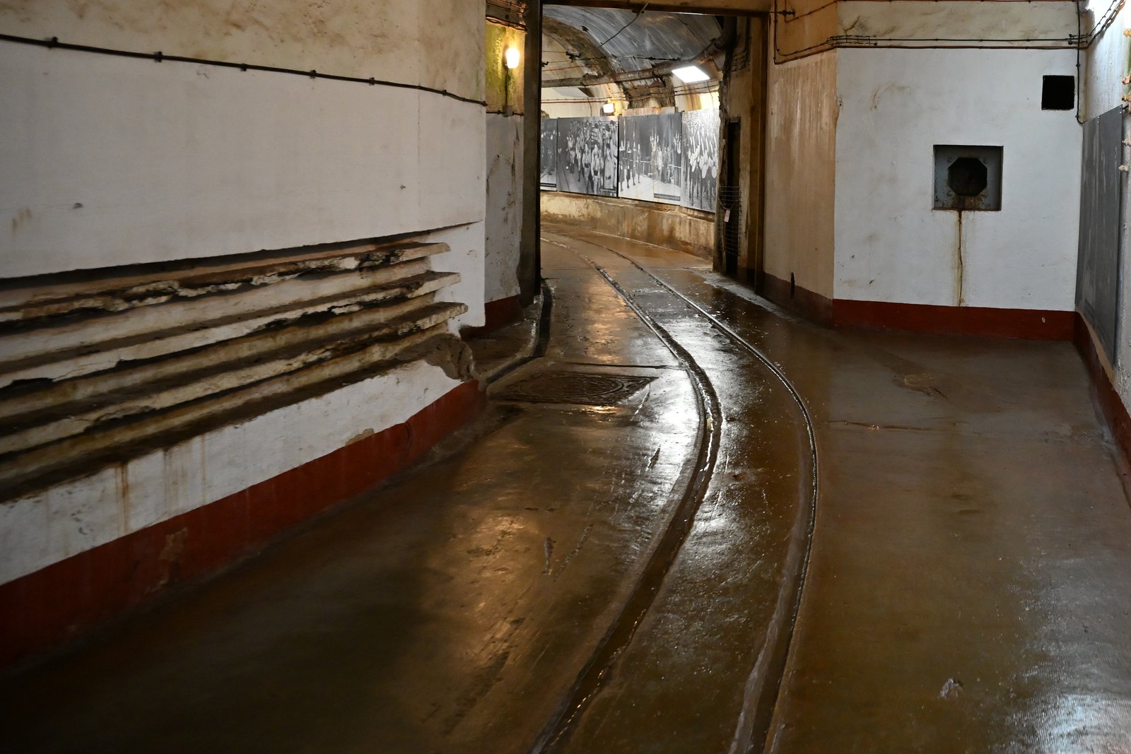 Galeria do forte de Hackenberg com o piso úmido, os trilhos fazendo uma curva e cabos correndo pelas paredes