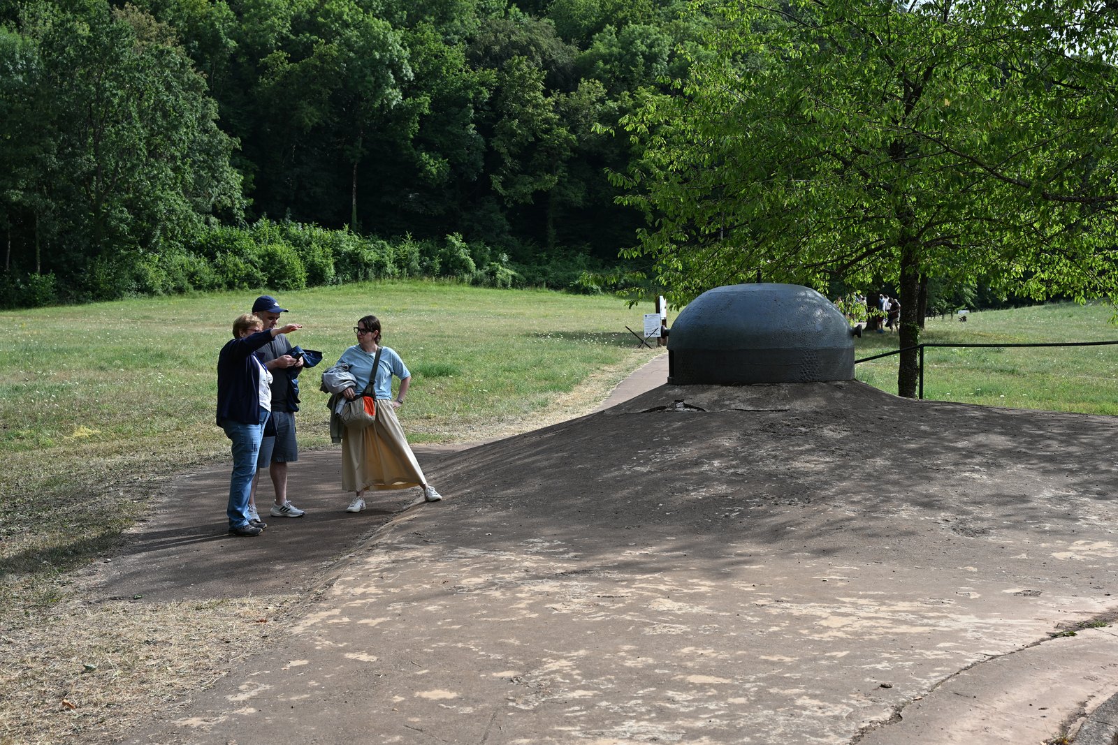 Três visitantes conversam ao lado de uma cloche blindada do Hackenberg, sobre a laje de concreto, com o prado e a mata ao fundo
