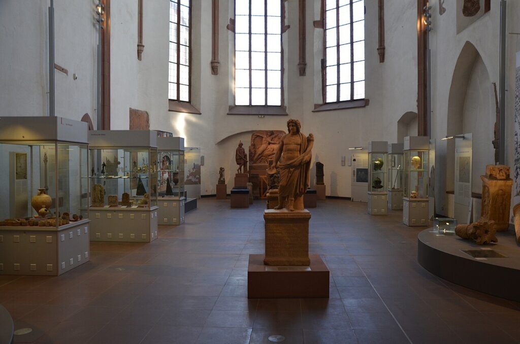 Salão de exposição do Museu Arqueológico de Frankfurt, com vitrines e uma estátua de pedra ao centro