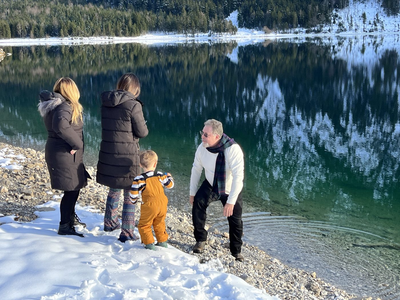 Família à beira de um lago alpino com neve, montanhas refletidas na água