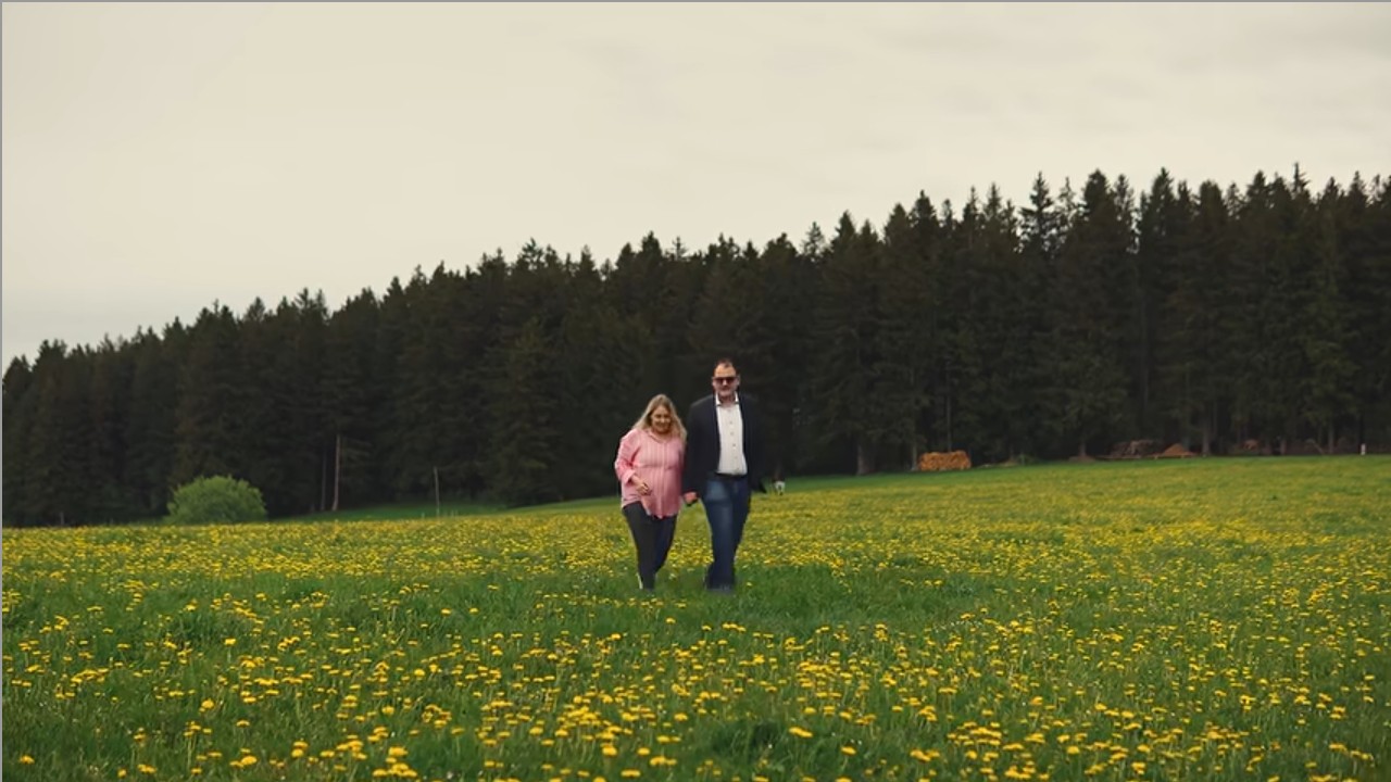 Os dois apresentadores caminhando juntos por um campo florido — cena do vídeo Quem somos