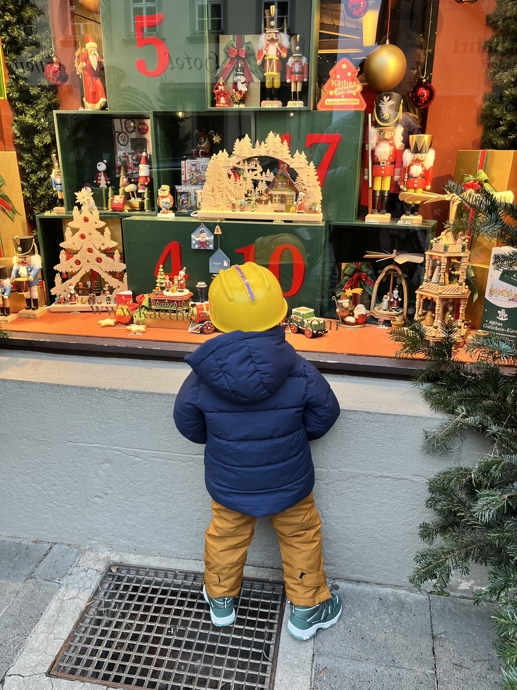 Niño con abrigo azul y casco amarillo mirando la vitrina de decoraciones navideñas de Käthe Wohlfahrt en Rothenburg ob der Tauber