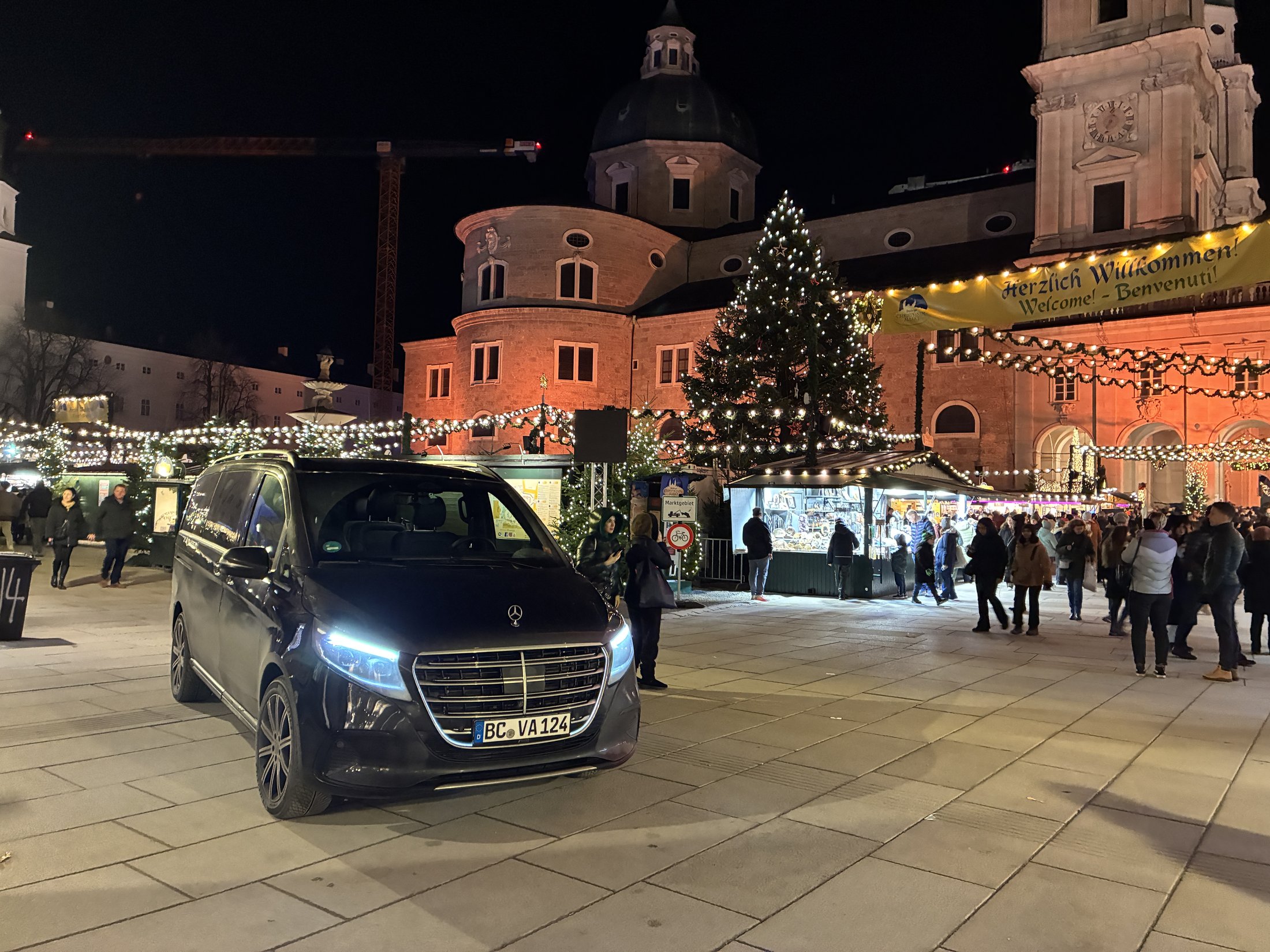 Mercedes Clase V negra de Viagem Alemanha estacionada en el mercado de Navidad de Salzburgo, con la catedral y el árbol de Navidad iluminados al fondo