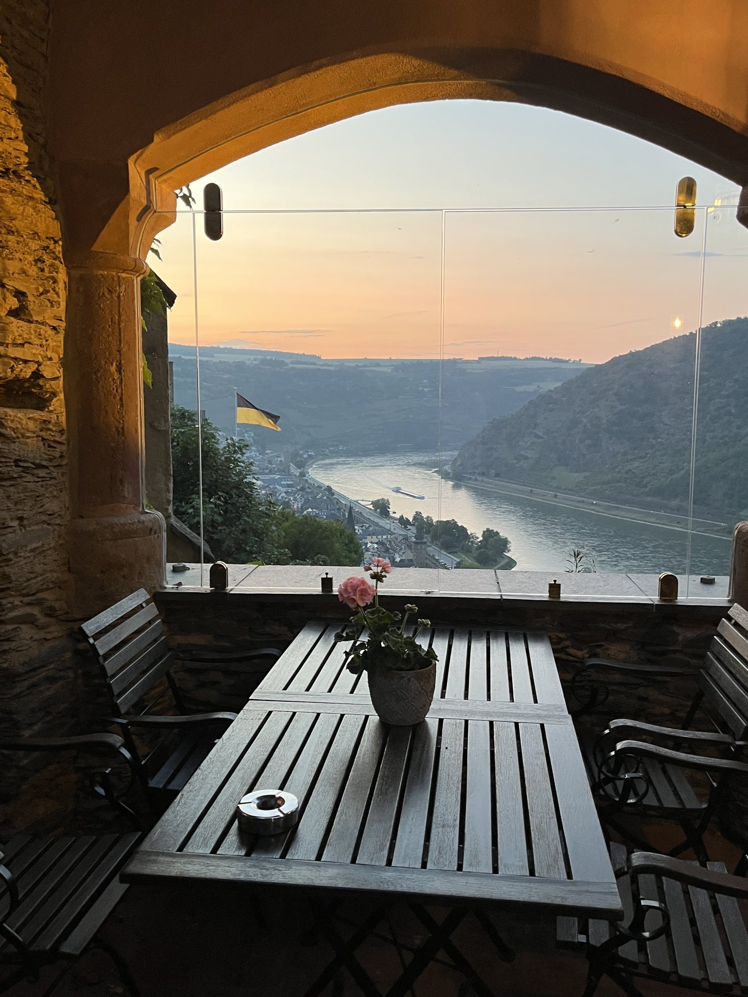 Terraço de castelo com mesa e vaso de flor, olhando o Reno ao pôr do sol, com a cidade de Oberwesel lá embaixo