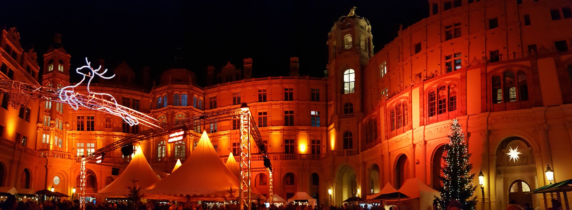 Panorámica nocturna del patio del castillo de Schwerin iluminado en naranja, con casetas del mercado de Navidad y un árbol de Navidad