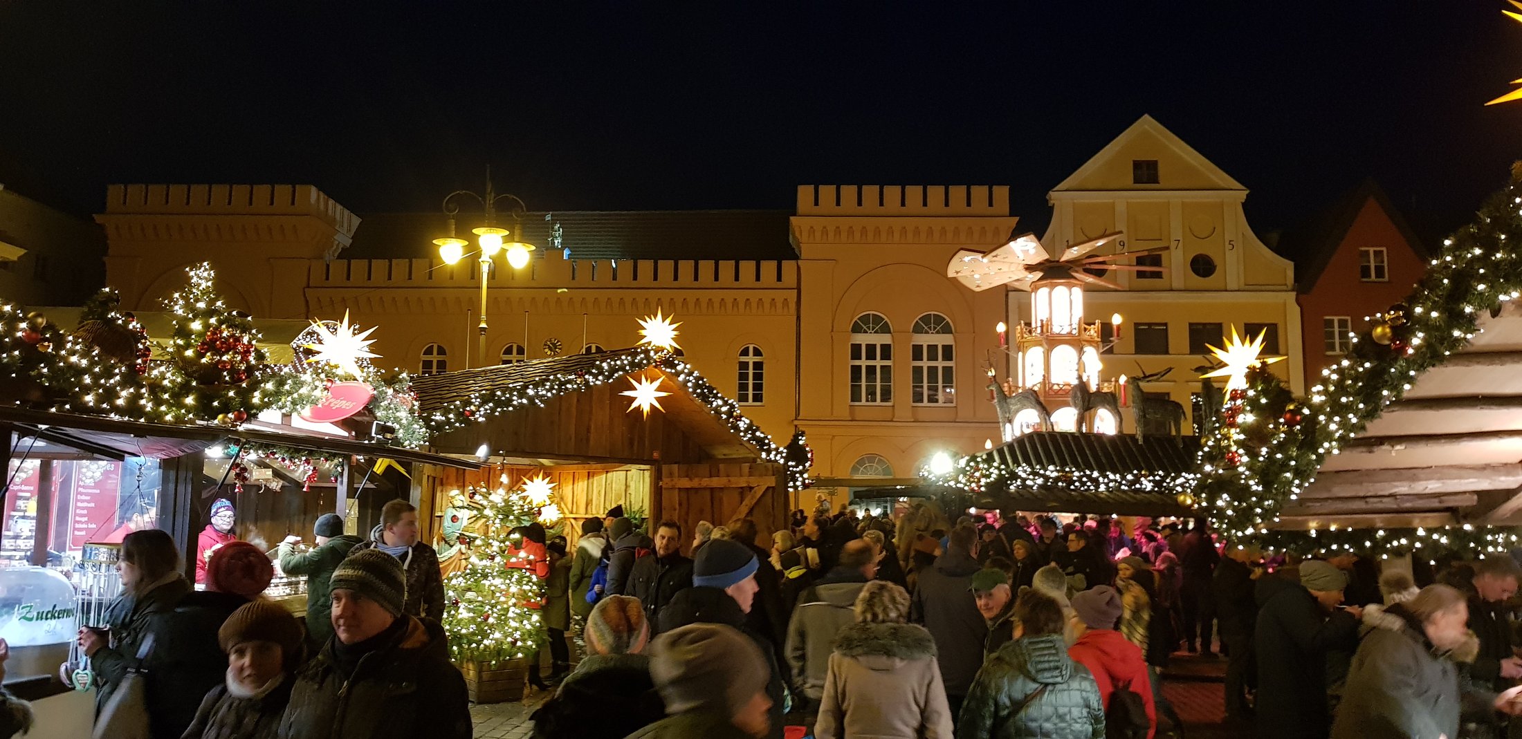 Mercado de Navidad de Schwerin de noche, con la pirámide navideña iluminada, estrellas de luz y el ayuntamiento amarillo al fondo