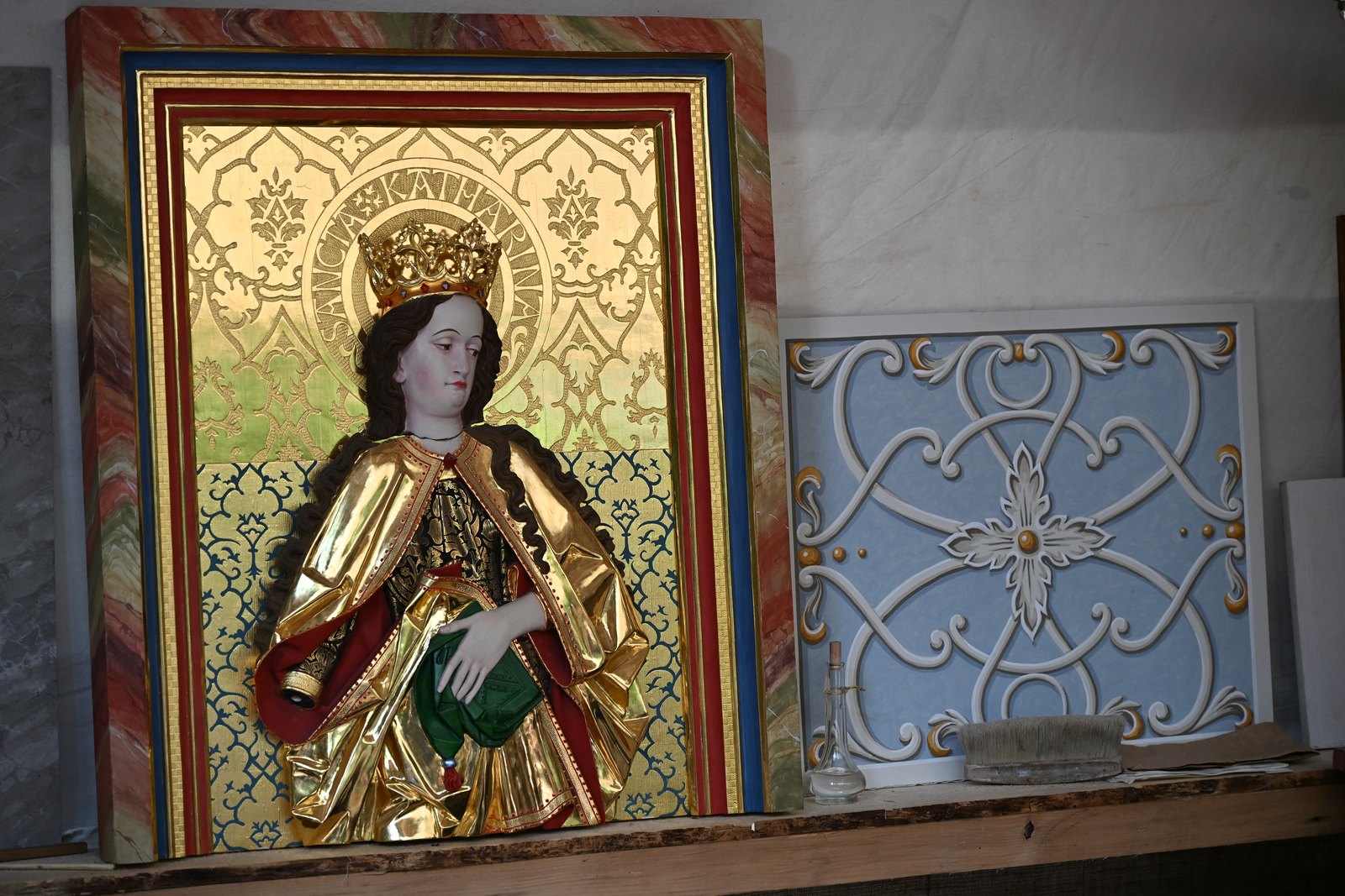 Painel de talha dourada com a inscrição SANCTA KATHARINA e figura coroada de manto dourado, ao lado de painel azul-claro com arabescos brancos, na banca de um dourador em Memmingen