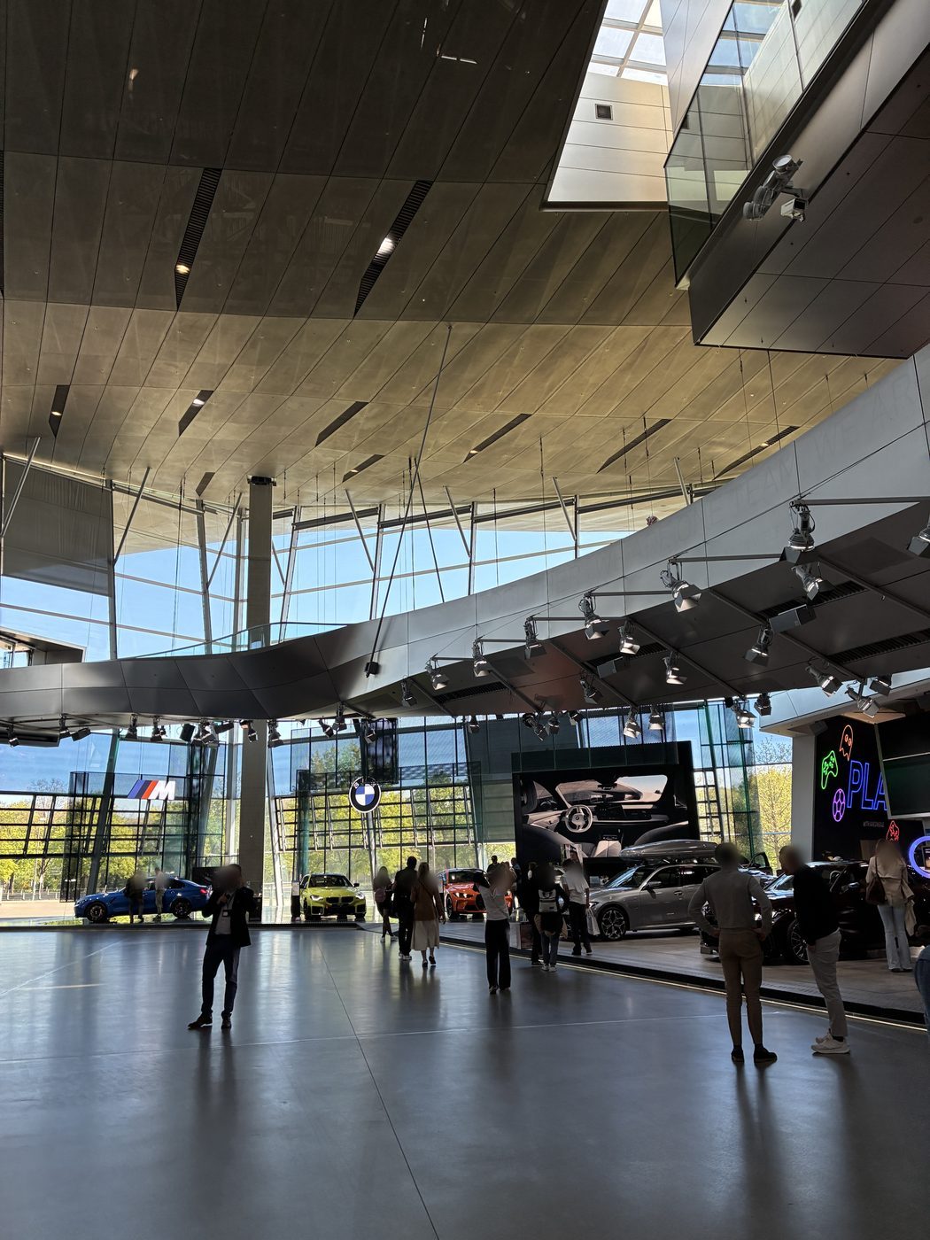 O salão do BMW Welt visto de baixo, com a cobertura ondulada, a passarela suspensa em curva, a área BMW M e carros em exposição