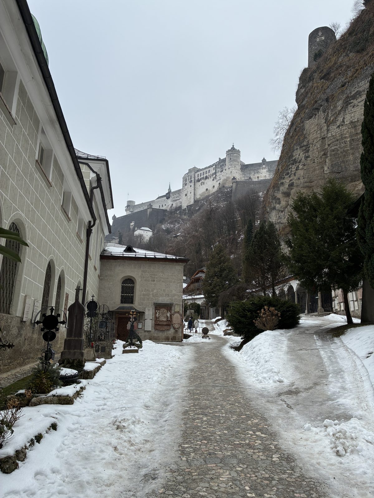 Caminho coberto de neve no cemitério de St. Peter, entre túmulos com grades de ferro e paredes de pedra, com a Fortaleza Hohensalzburg no alto do rochedo