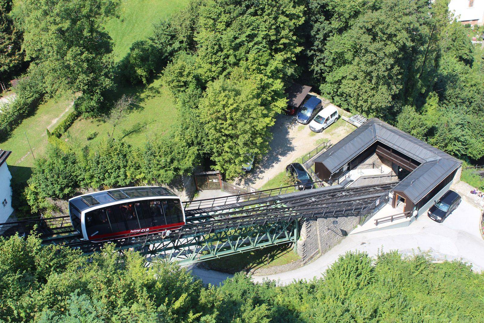 Carro vermelho e branco da FestungsBahn sobre a ponte de ferro do trilho, visto de cima, entre árvores e o telhado da estação