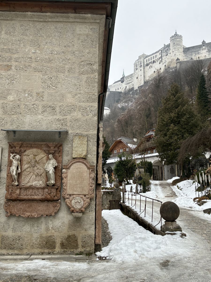 Viela de St. Peter coberta de neve, com lápides na parede de pedra à esquerda e a fortaleza branca no alto do morro ao fundo