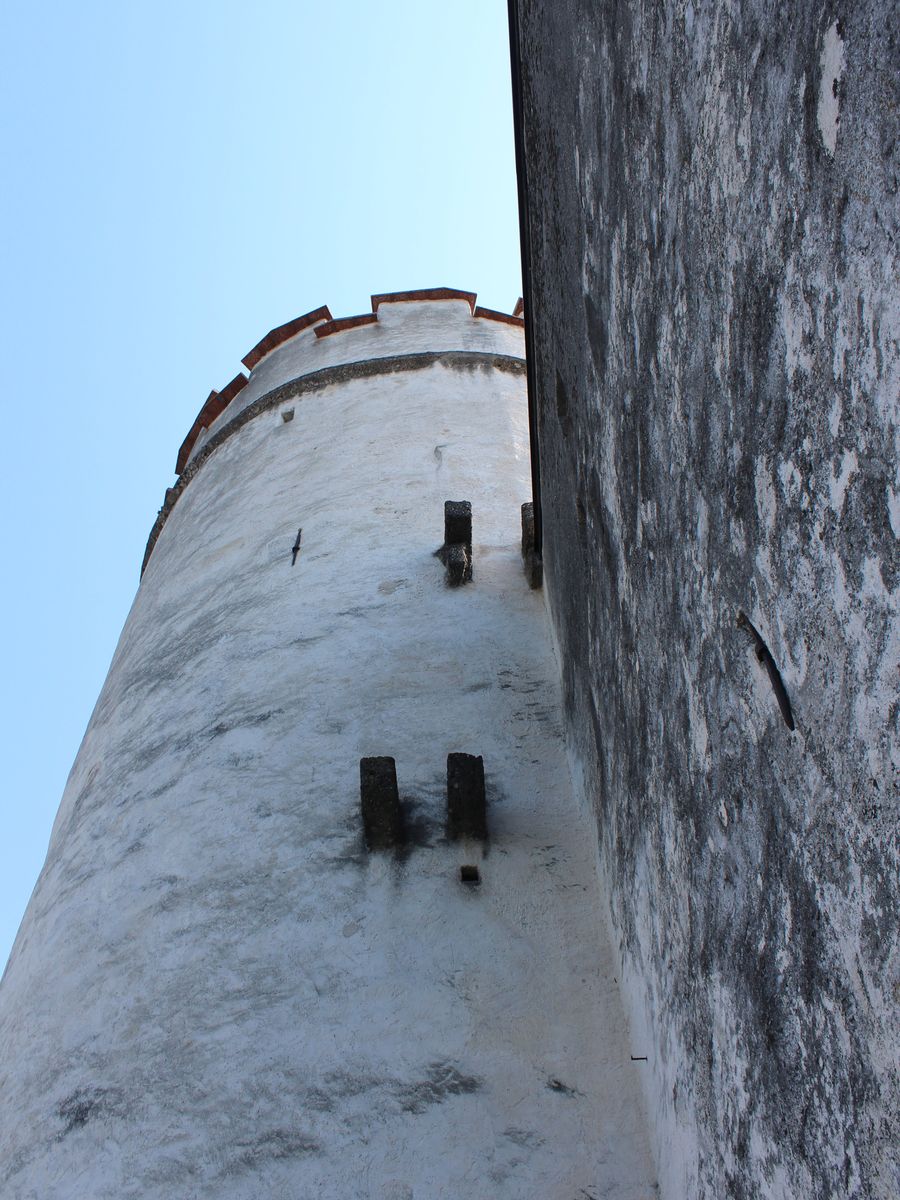 Torre redonda e caiada da Fortaleza Hohensalzburg, com ameias no alto, vista de baixo contra o céu azul, ao lado de uma muralha de pedra