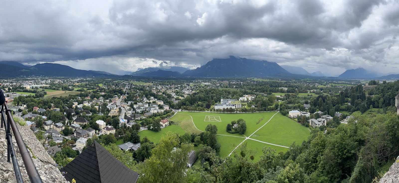 Panorama do alto da fortaleza para o sul de Salzburgo, com um grande campo verde, bairros arborizados e montanhas sob nuvens carregadas