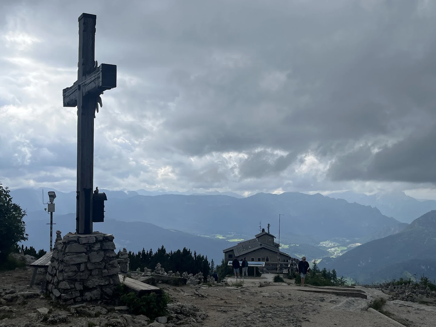 Cruz do cume do Kehlstein em primeiro plano, com caminho de pedra, o Kehlsteinhaus abaixo e montanhas sob nuvens