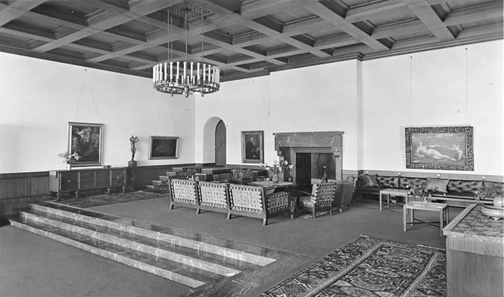 Grande salão vazio do Berghof, com sofás, lareira, quadros e degraus