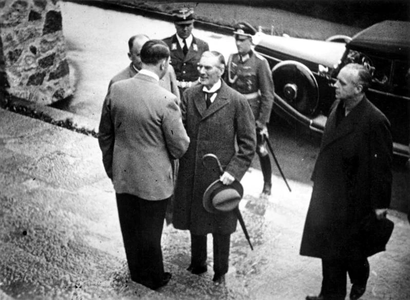 Hitler de costas cumprimenta Neville Chamberlain, com Ribbentrop à direita e um carro ao fundo