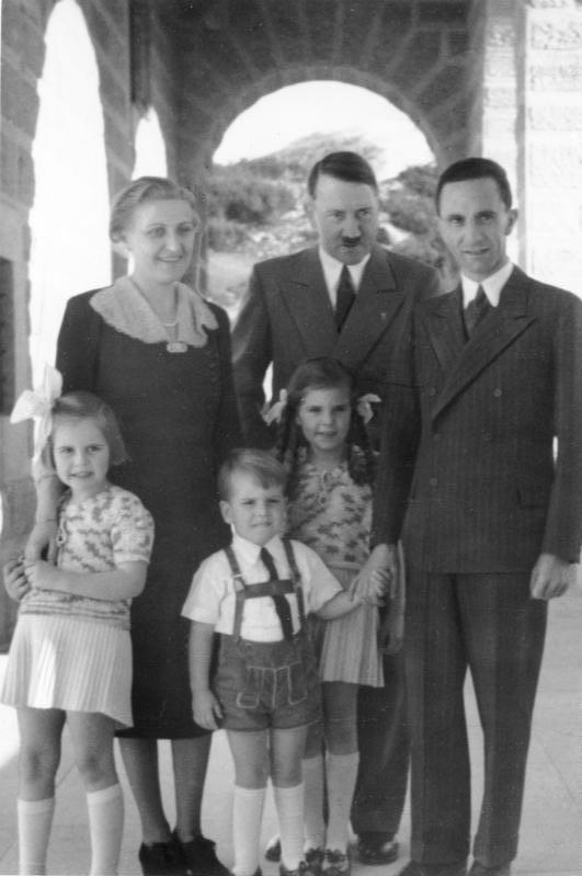 Fotografia em preto e branco de Hitler, Joseph e Magda Goebbels e três crianças, todos em pé numa arcada de pedra aberta, com montanhas ao fundo