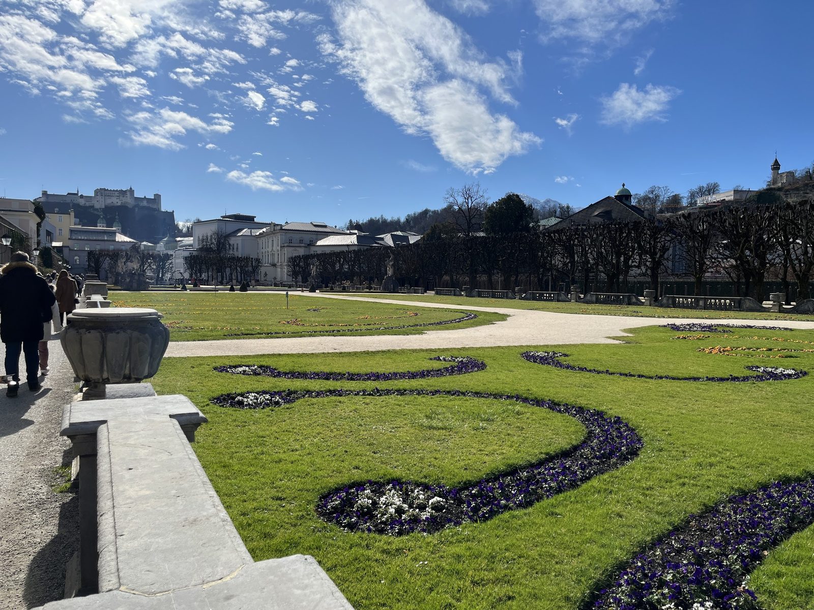 Canteiros desenhados dos Jardins de Mirabell com balaustrada de pedra e a Fortaleza Hohensalzburg ao fundo, sob céu azul com nuvens