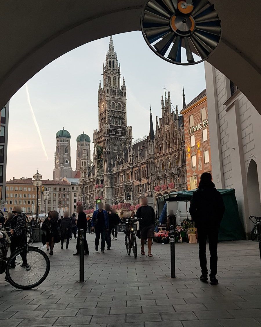 A Marienplatz vista através de uma passagem em arco, com a torre neogótica do Neues Rathaus, as duas torres de cúpula verde da Frauenkirche ao fundo e pedestres na praça ao entardecer