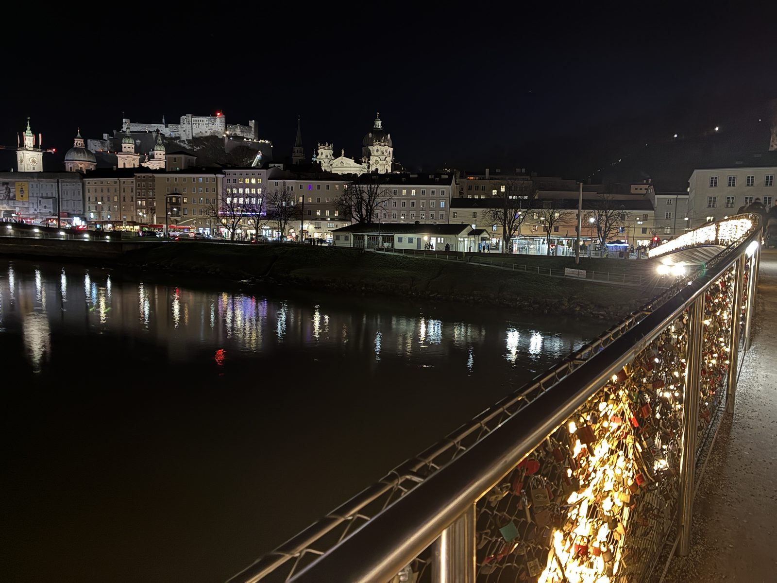 Grade da ponte Marko-Feingold-Steg coberta de cadeados, com o rio Salzach escuro e a fortaleza iluminada ao fundo, à noite