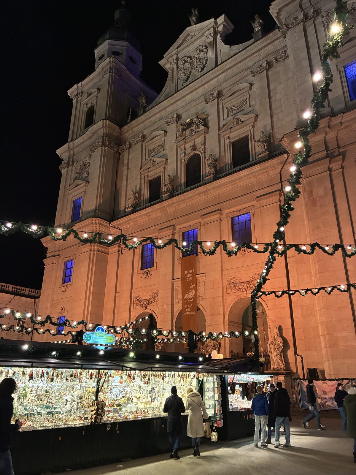 Mercado de Natal diante da fachada iluminada da Catedral de Salzburgo à noite, com barracas de enfeites e guirlandas de luzes