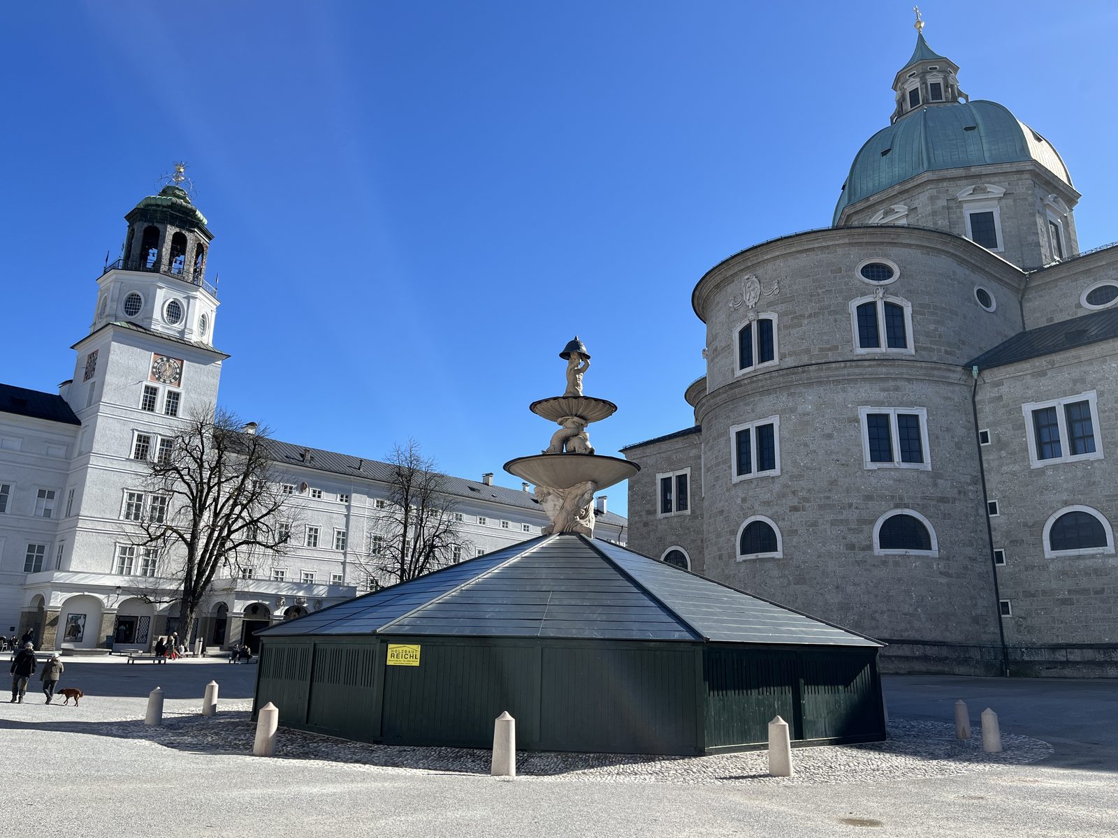 Residenzplatz no inverno, com o chafariz protegido por uma cobertura escura, a catedral à direita e a torre do Glockenspiel à esquerda