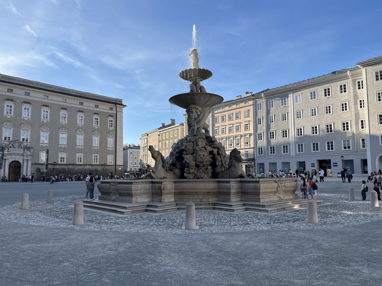 Residenzbrunnen, chafariz barroco de várias bacias com cavalos esculpidos, no meio da Residenzplatz, com os prédios da Residência ao redor, numa tarde de abril