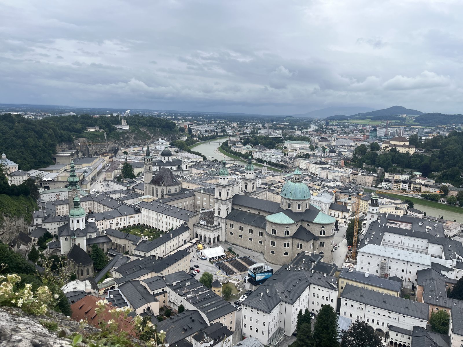 Vista do alto da Fortaleza Hohensalzburg sobre o centro histórico de Salzburgo, com a cúpula verde da catedral, praças, o rio Salzach ao fundo e morros arborizados dos dois lados, num dia nublado de verão