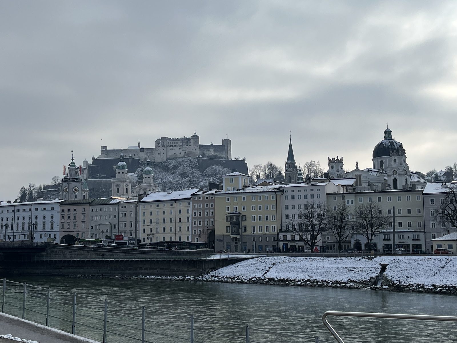 A Cidade Velha de Salzburgo com telhados e margens do rio Salzach cobertos de neve e a Fortaleza Hohensalzburg no alto, num dia nublado de inverno
