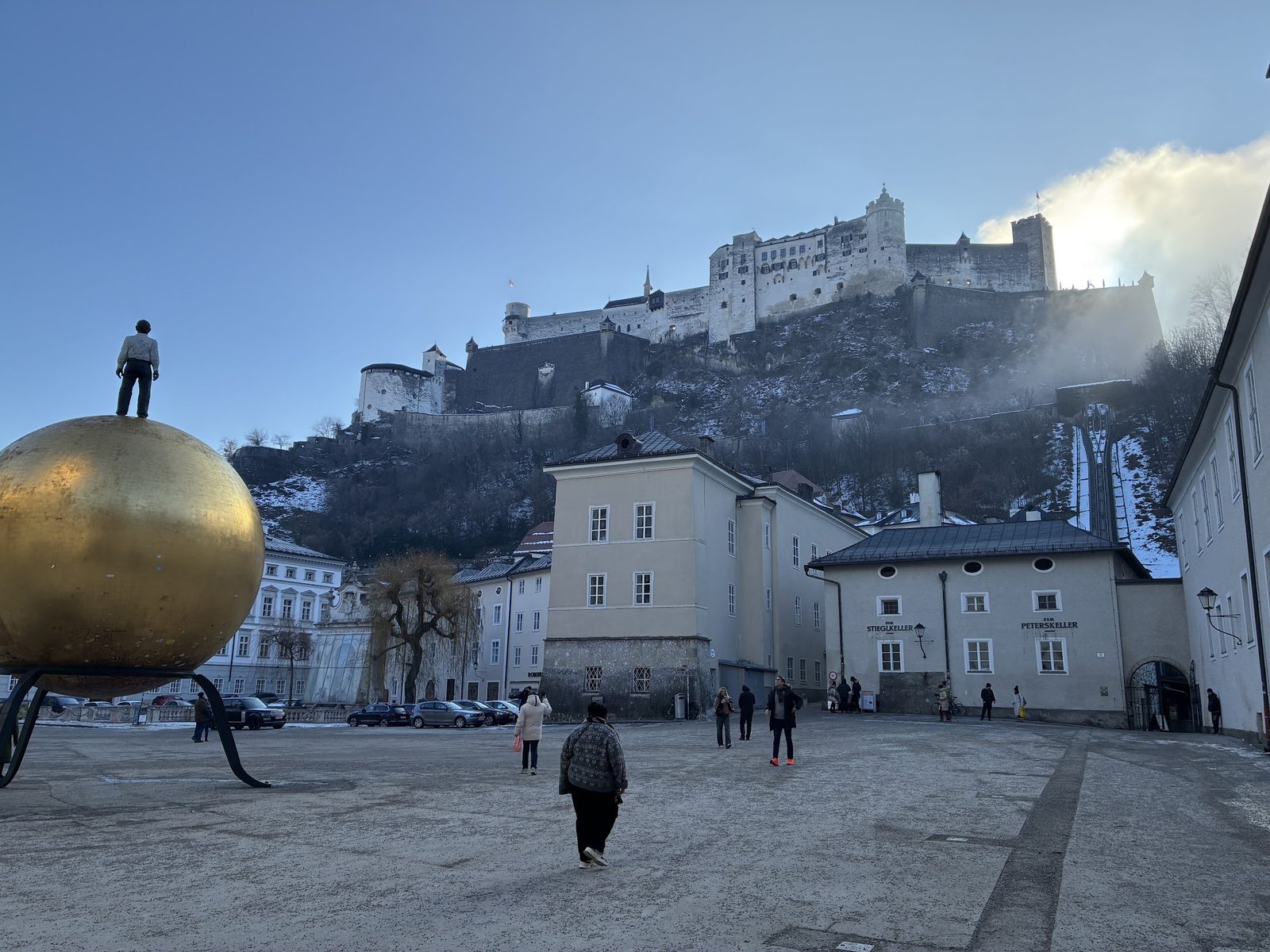 A Sphaera, grande esfera dourada com a escultura de um homem em cima, no Kapitelplatz, com a Fortaleza Hohensalzburg no alto numa manhã de inverno