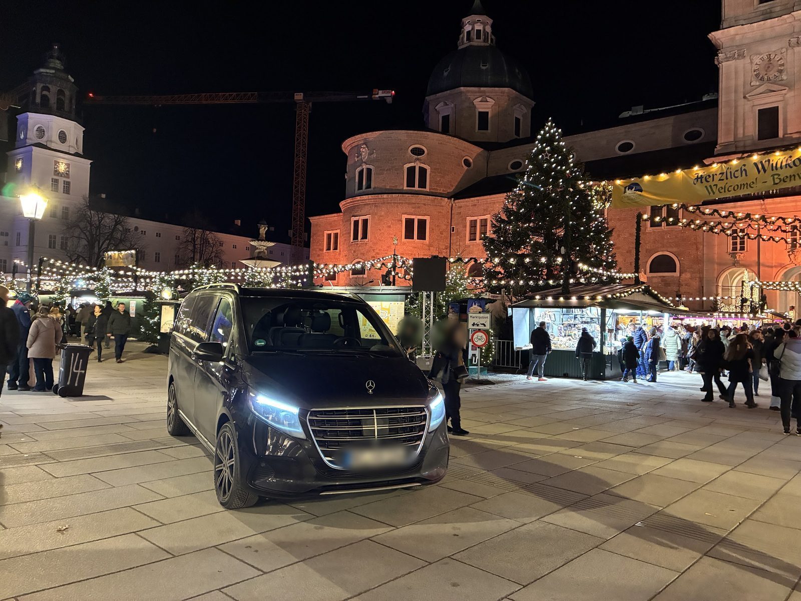 Van preta de passageiros estacionada na Domplatz diante do mercado de Natal iluminado e da catedral, à noite
