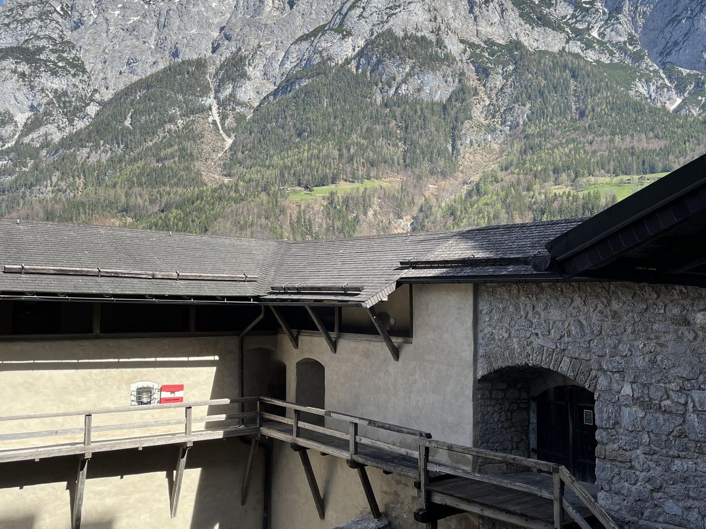 Pátio interno do Castelo de Hohenwerfen, com passarelas de madeira e muros caiados, e as montanhas rochosas do Tennengebirge com neve ao fundo