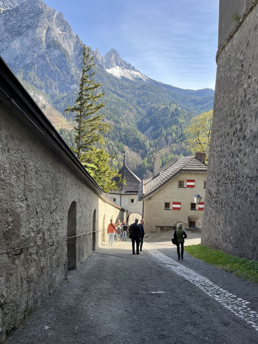 Rua de paralelepípedo entre muralhas altas dentro do Castelo de Hohenwerfen, com visitantes de costas e o paredão do Tennengebirge ao fundo