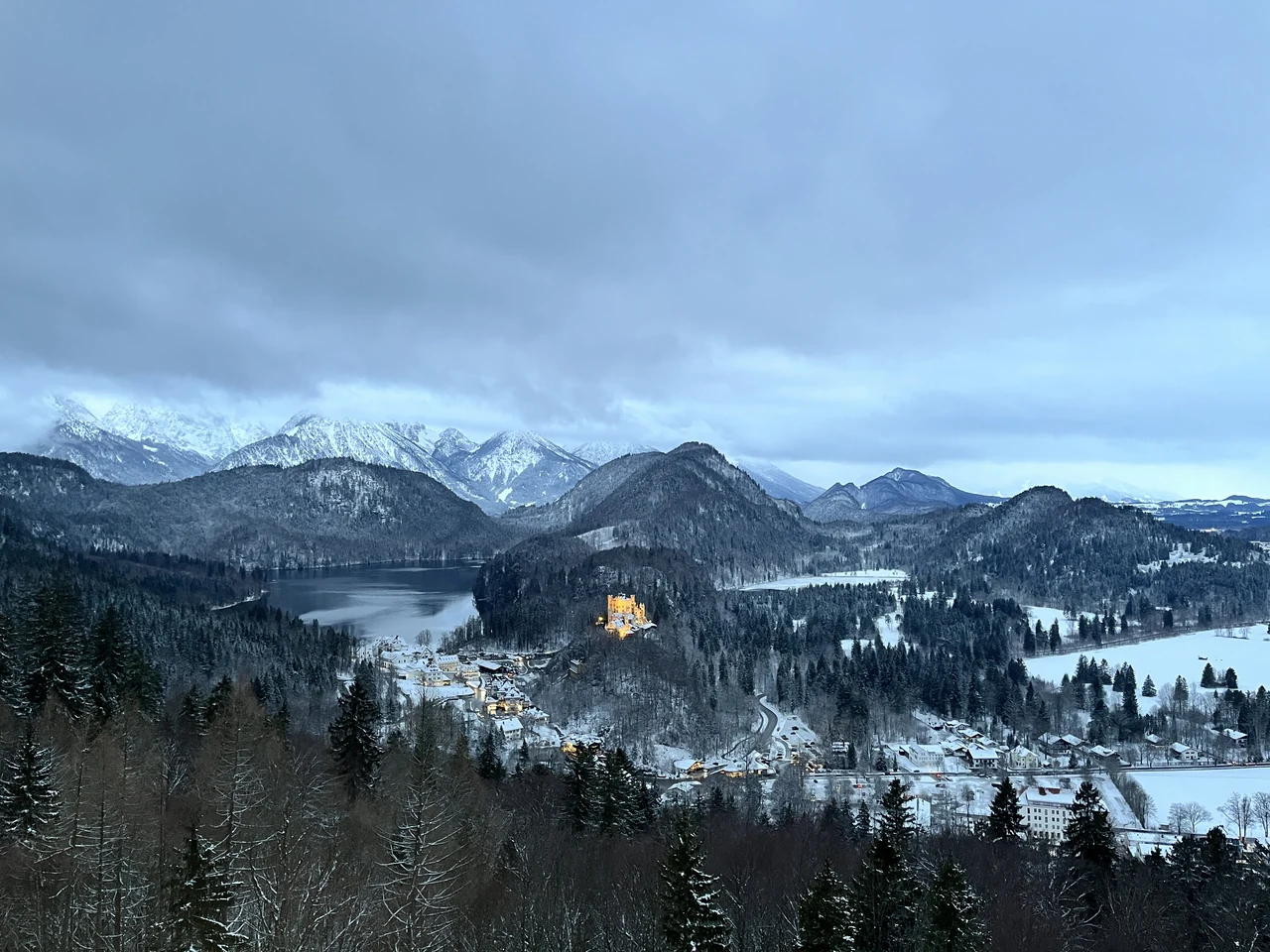 Castelo de Hohenschwangau iluminado diante do Alpsee e de montanhas com neve ao entardecer