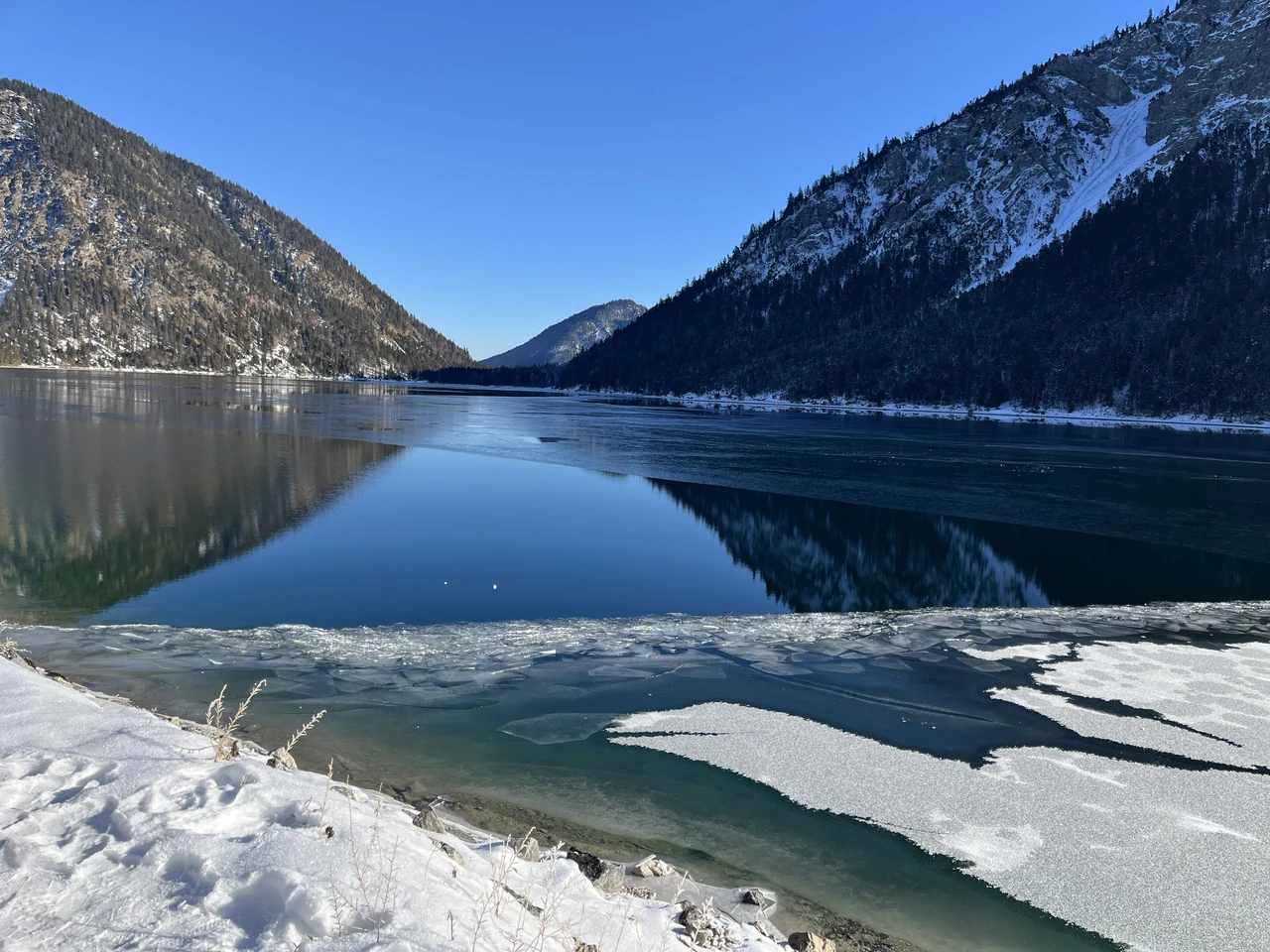 Montanhas e árvores refletidas no Plansee, com gelo e neve junto à margem