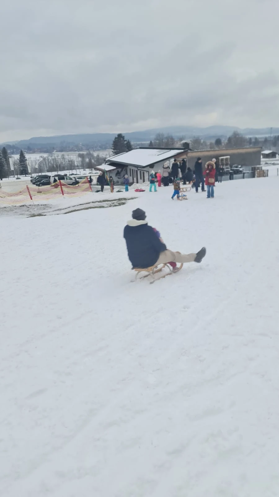 Pessoa de costas descendo uma encosta de neve em um trenó, com a base da pista ao fundo