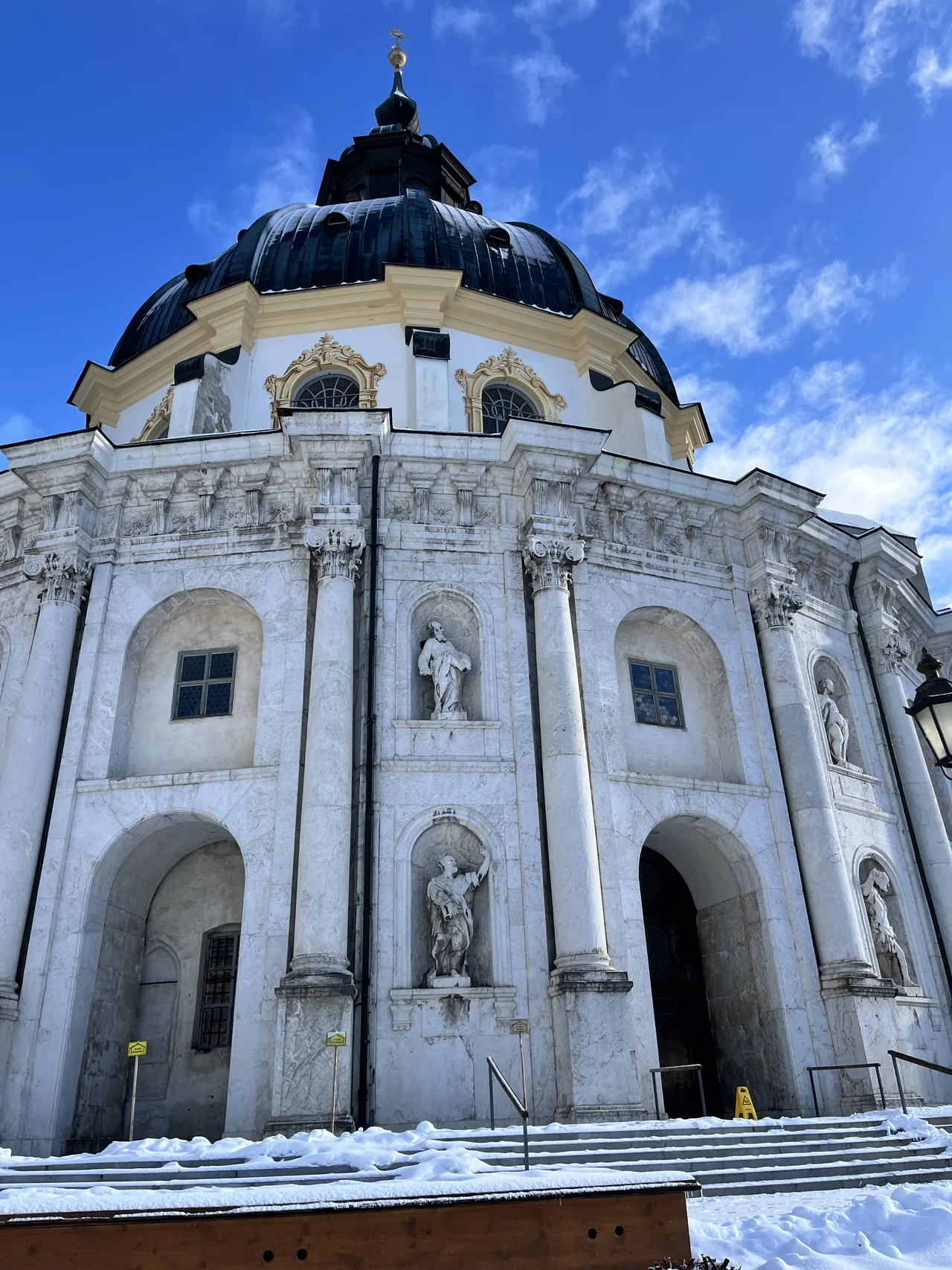 Fachada da Abadia de Ettal sob céu azul, com neve nos degraus em dezembro