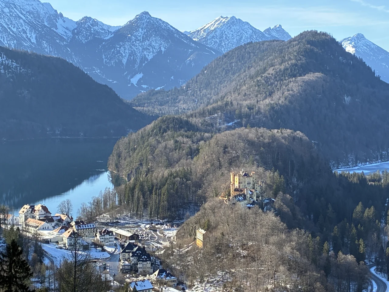 Lago Alpsee à esquerda, vila de Hohenschwangau abaixo e palácio amarelo na encosta, com neve parcial e montanhas ao fundo
