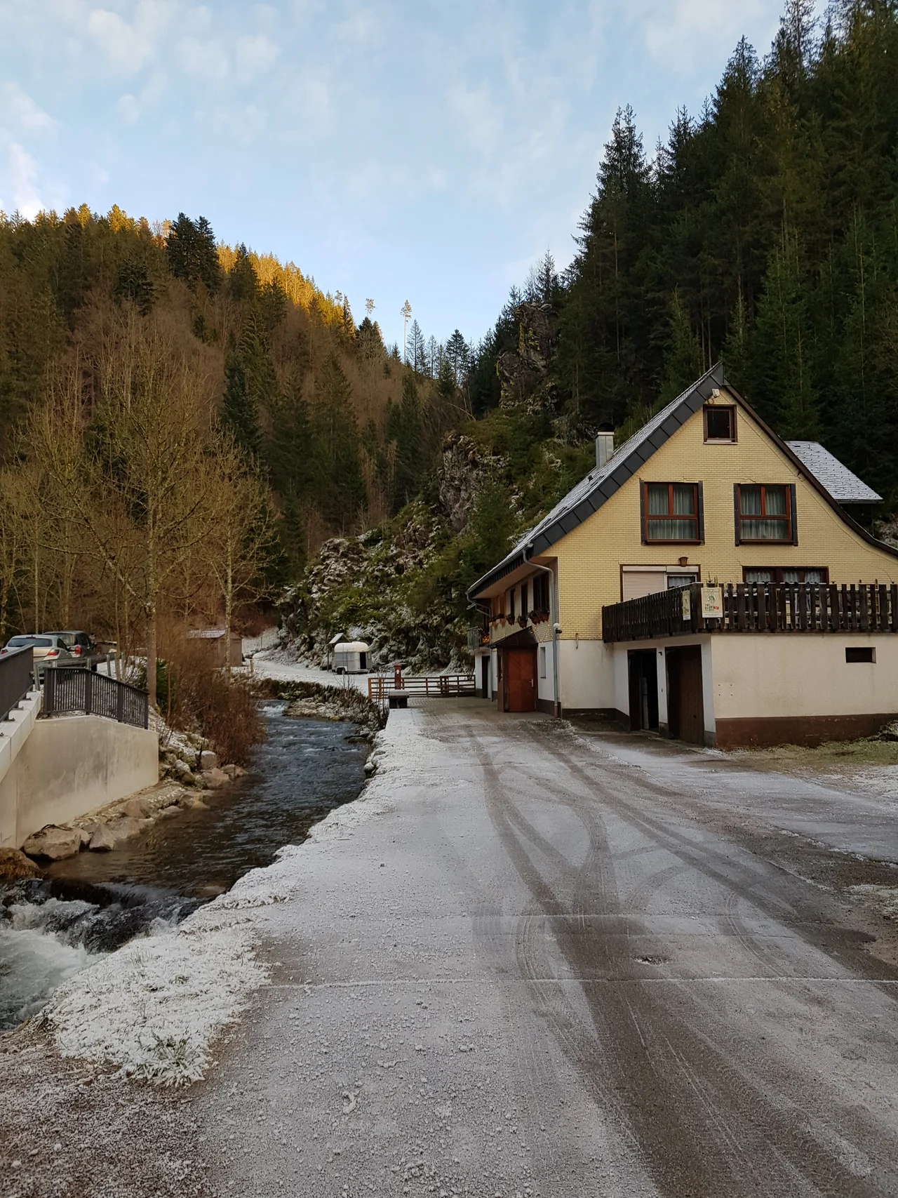 Rio correndo junto a uma casa e árvores na Floresta Negra, com neve nas margens