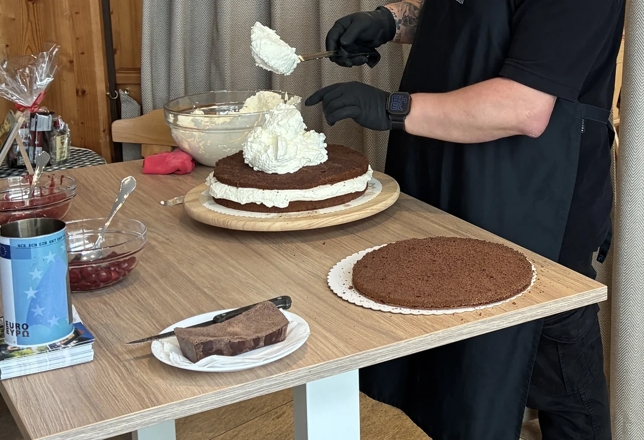 Mãos acrescentando creme às camadas de uma torta Floresta Negra, com cerejas e chocolate sobre a mesa